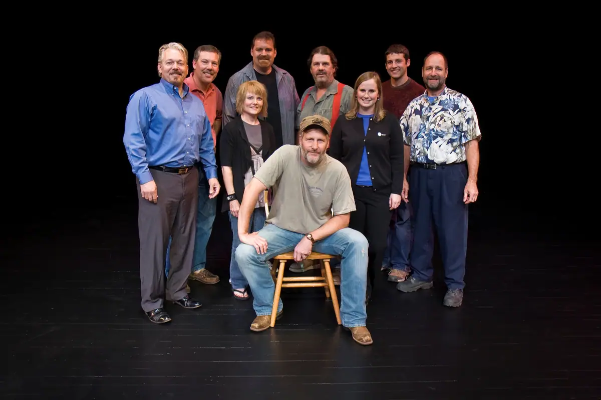 Nine adults pose together on a dark stage, with one person seated on a wooden stool.