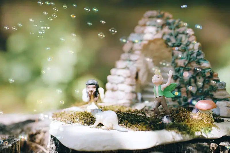 A miniature fantasy garden with winged figures, a white mouse, bubbles, moss, and a stone arch.