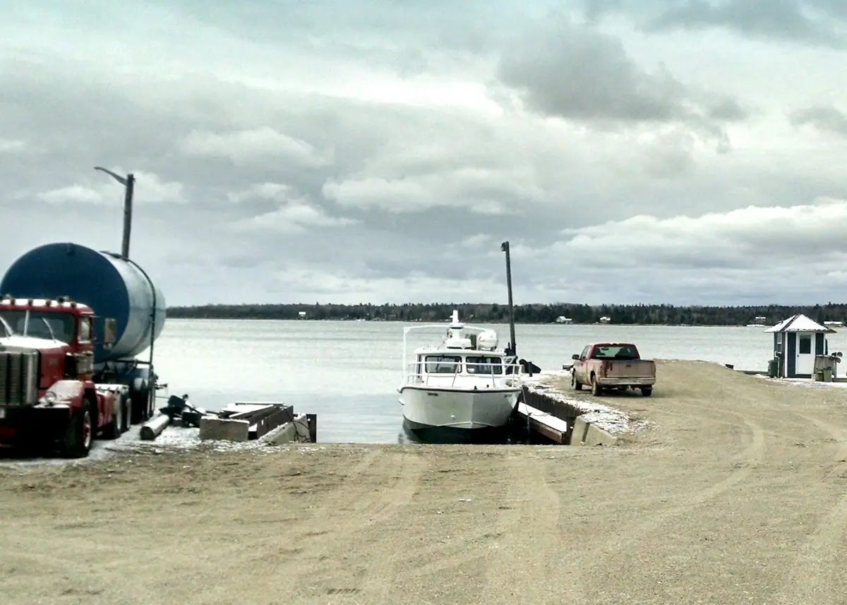 A white cabin boat at a harbor ramp beside a tanker truck and pickup under a cloudy sky.