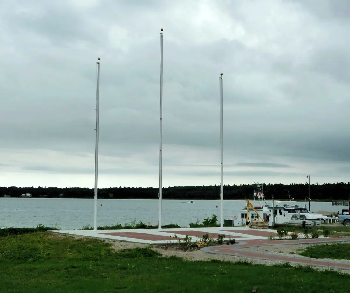 Three empty flagpoles rise above a patterned waterfront plaza beside moored boats.