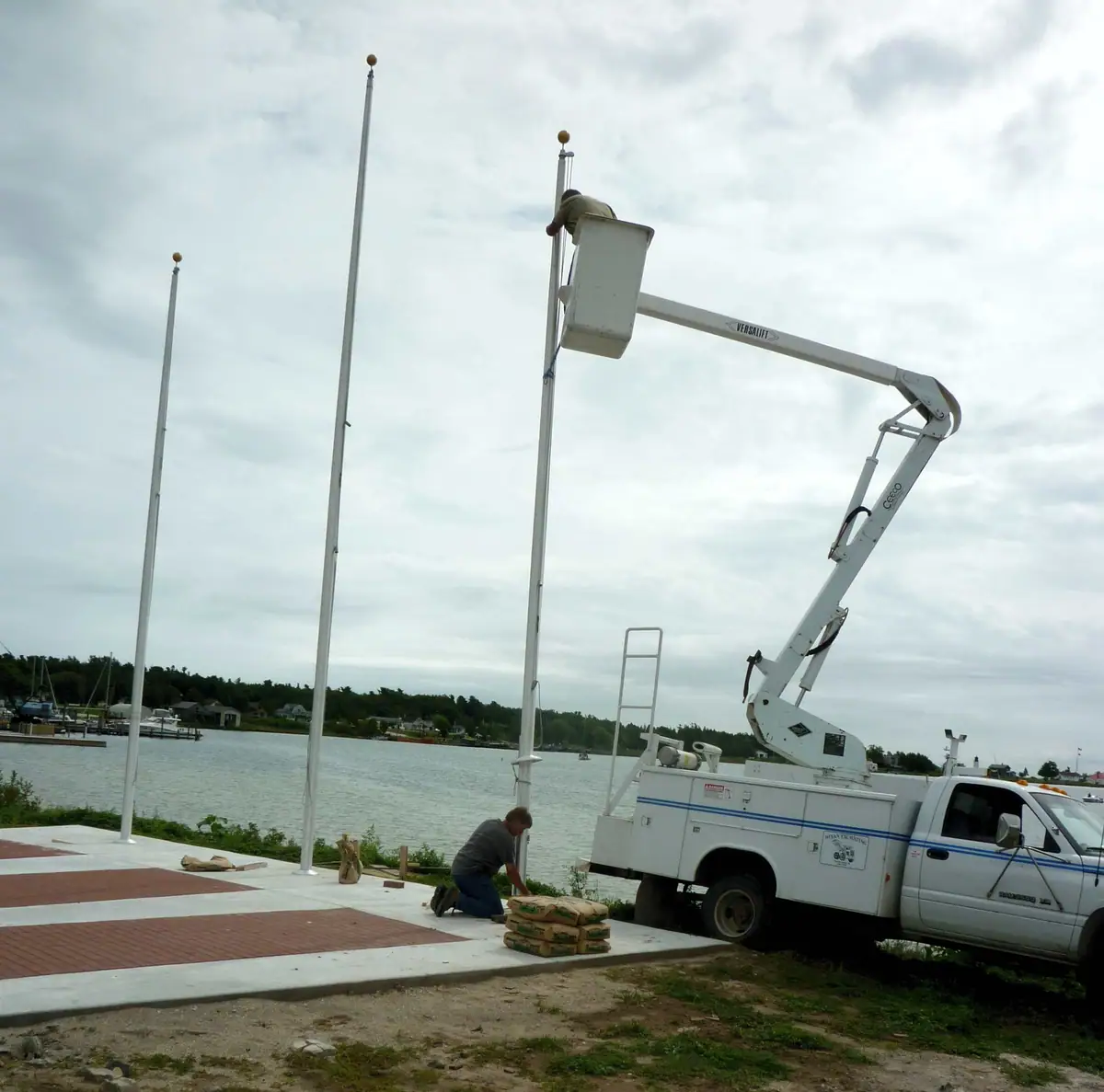 A worker uses a bucket truck to service a flagpole beside a waterfront promenade.