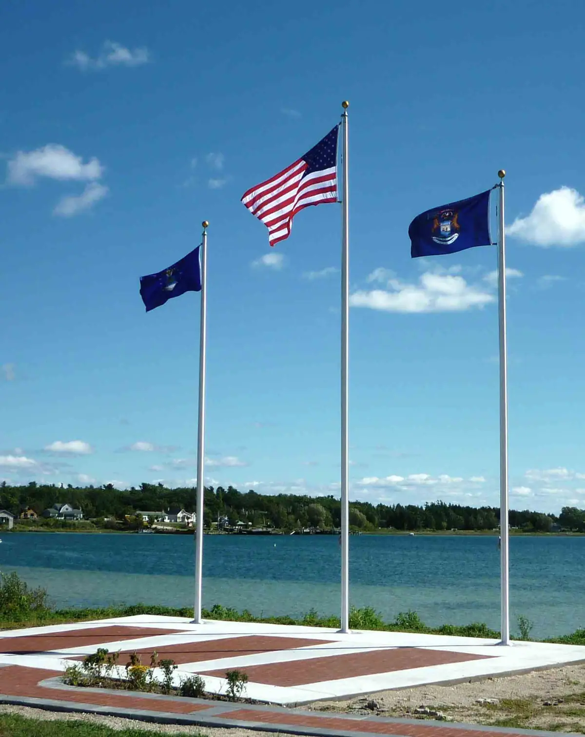 Three flags fly above a paved waterfront area, with houses and a wooded shoreline beyond the water.