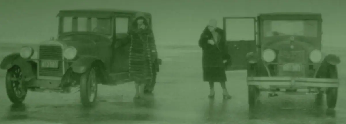 Two women pose beside two vintage automobiles on a broad, flat open surface.