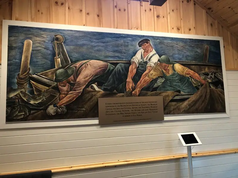 A framed painting shows three fishermen hauling a net aboard a small boat, with a dedication plaque below.
