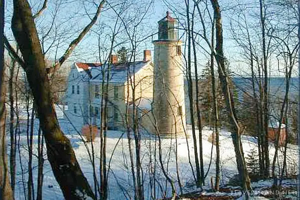 Snow-covered lighthouse and adjoining house stand among leafless trees beside visible water.
