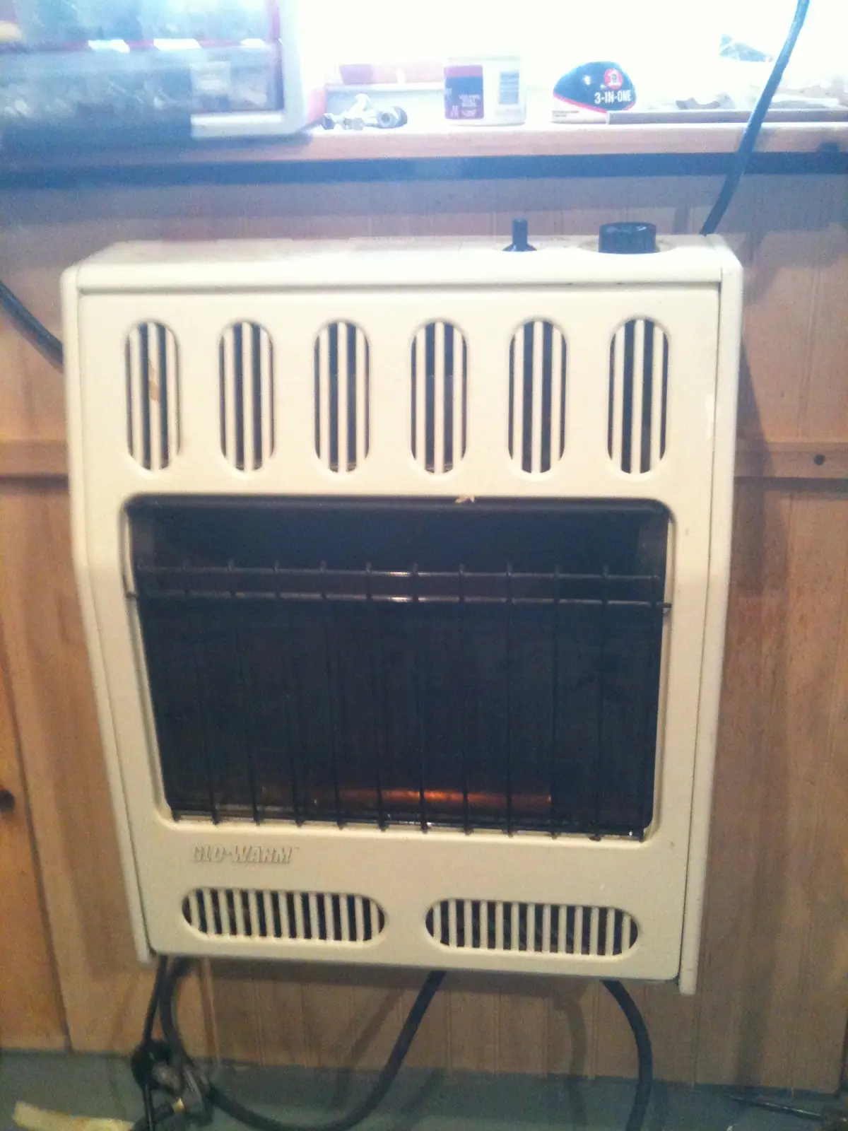 A white portable electric heater with a dark grille and glowing heating element stands against wood paneling.