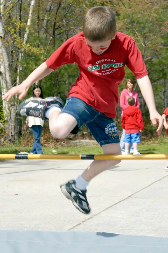 A child jumps over a yellow-and-black bar while others watch in a wooded outdoor setting.