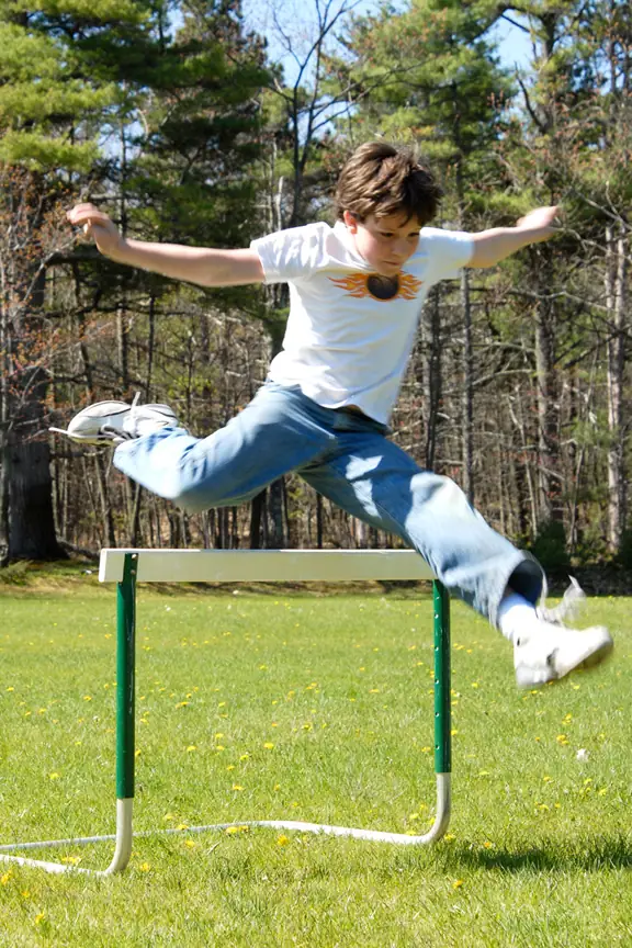 Young athlete clears a hurdle on a grassy field bordered by trees.