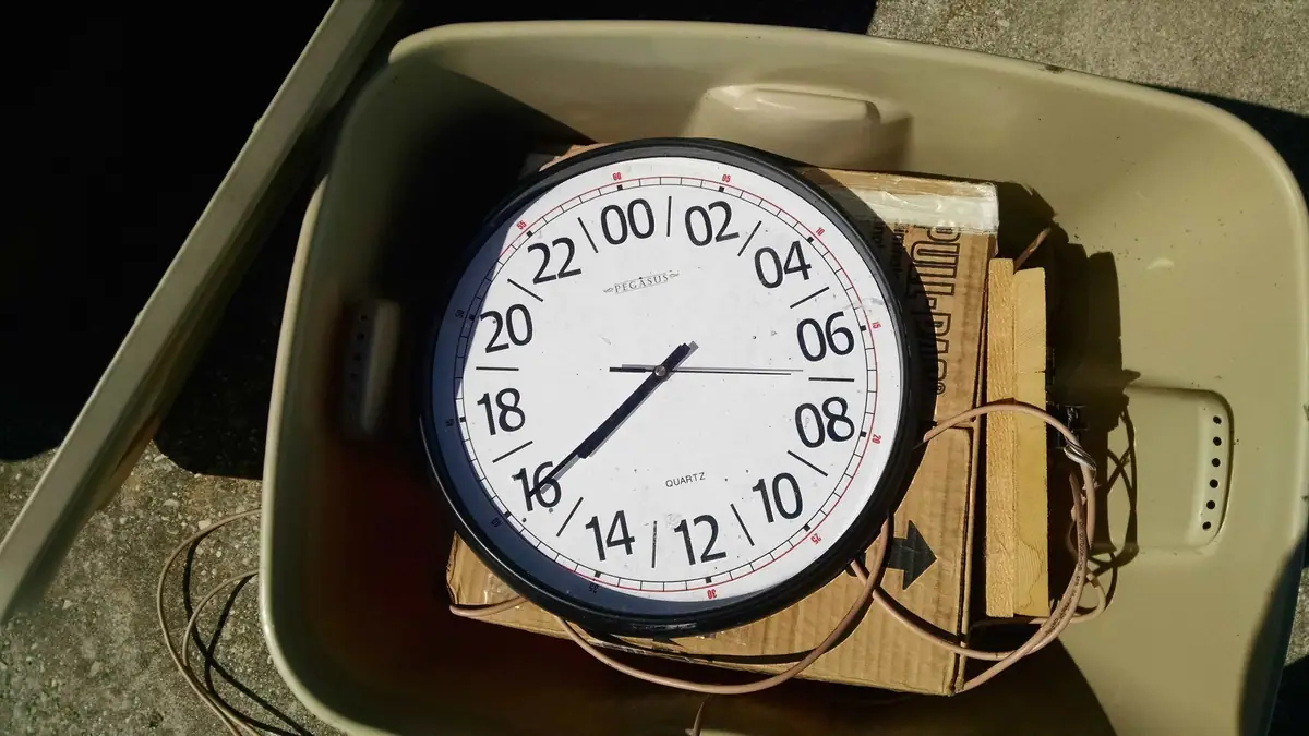 A round 24-hour clock rests on a cardboard box inside a tan plastic storage bin.