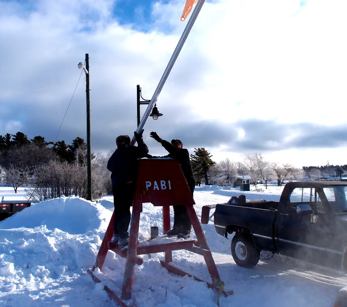 Two people work from a red ladder beside a streetlight in deep snow, with a pickup truck nearby.