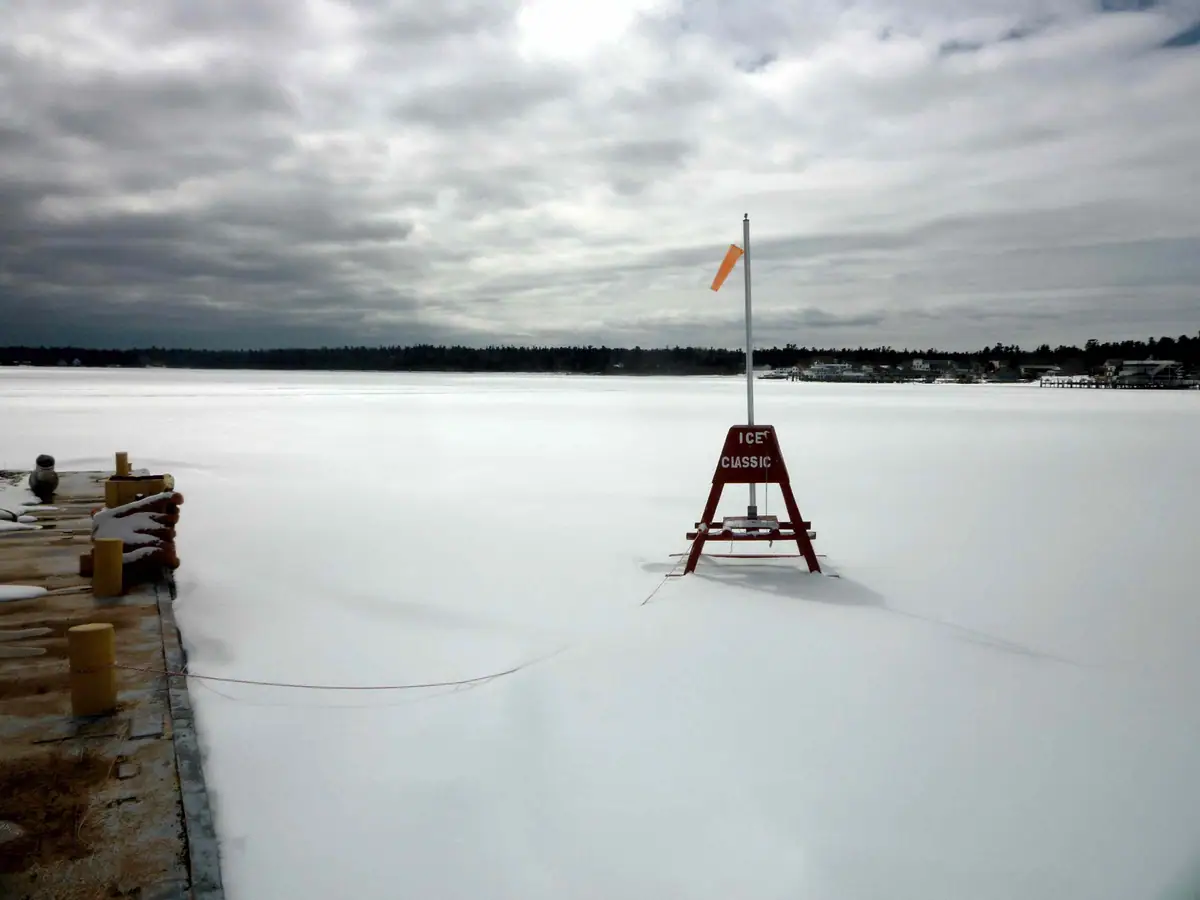 A red ICE CLASSIC marker and windsock stand on a frozen body of water beside a dock.