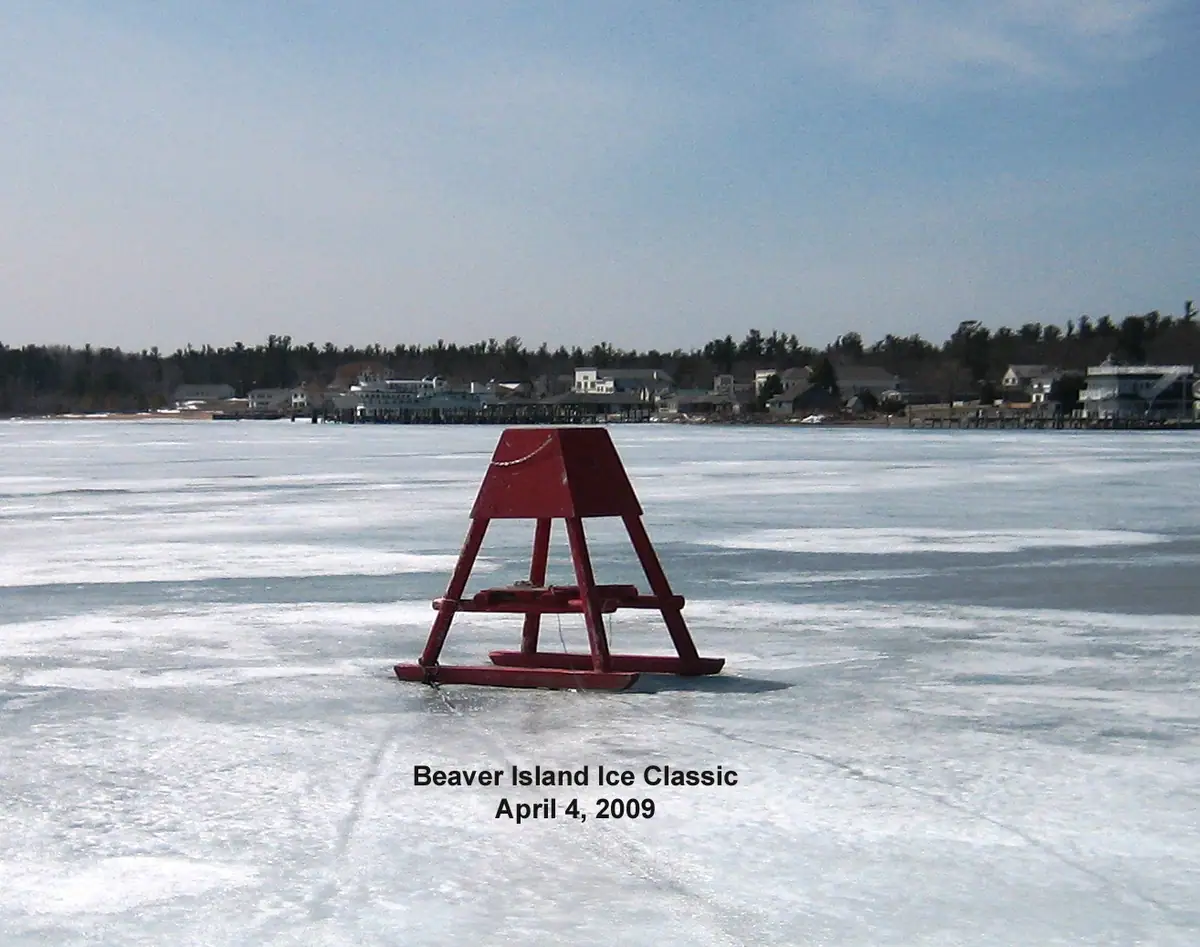 Red A-frame marker standing on frozen water, with houses and trees along the distant shore.