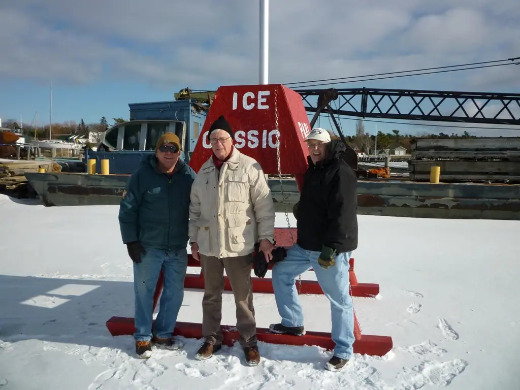 Three men pose beside a red ice-themed structure on a snow-covered waterfront with boats and a crane behind them.