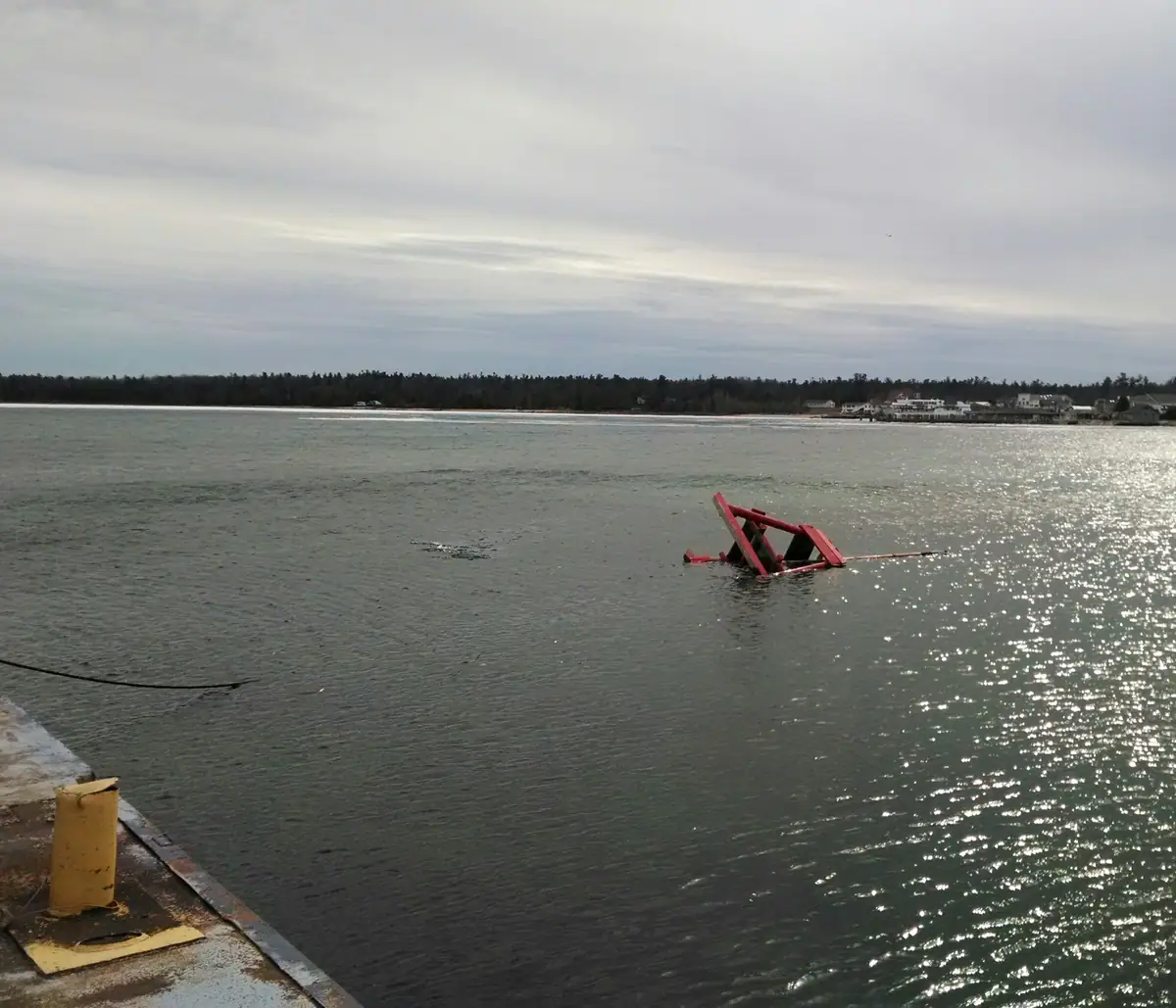 A red partially submerged structure floats near a concrete pier, with houses and trees along the far shoreline.