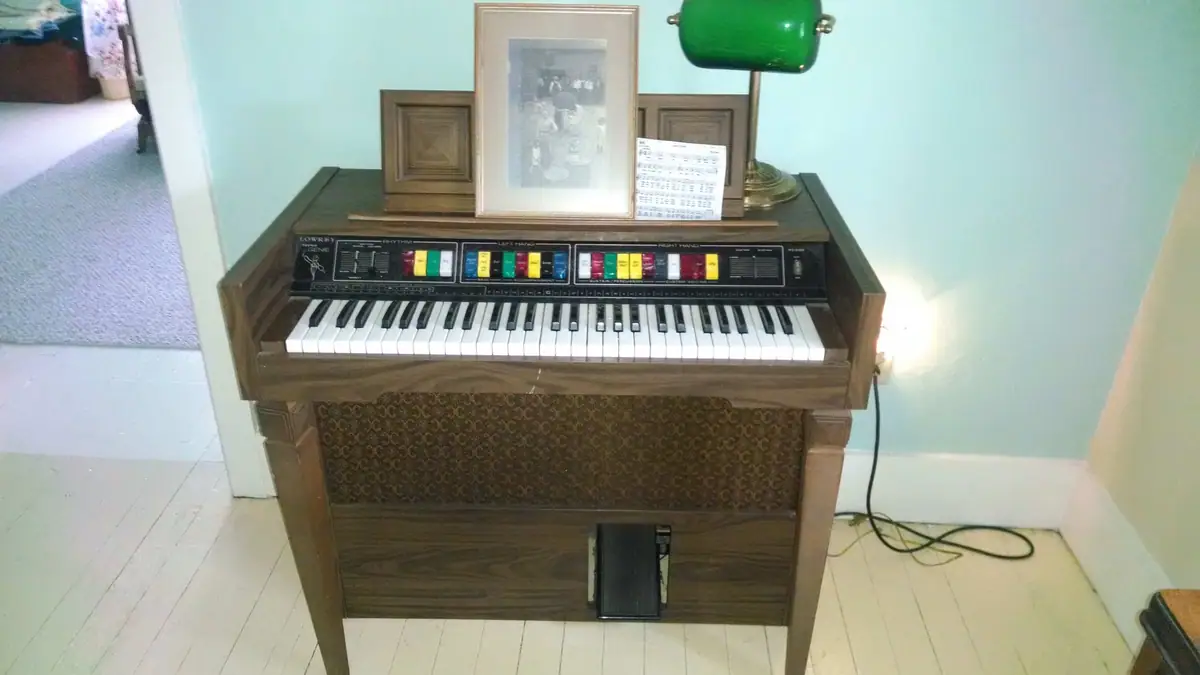 A wooden electronic organ with a keyboard and foot pedal stands against a wall beside framed pictures and a lamp.