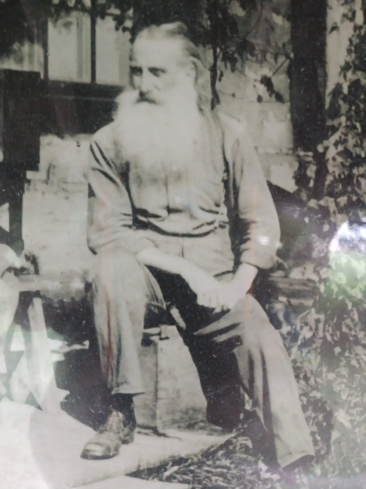 A bearded man sits in a chair beside a window and leafy plants in an aged monochrome portrait.