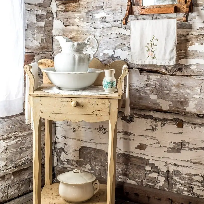 A vintage washstand with ceramic vessels stands against a peeling wooden wall.