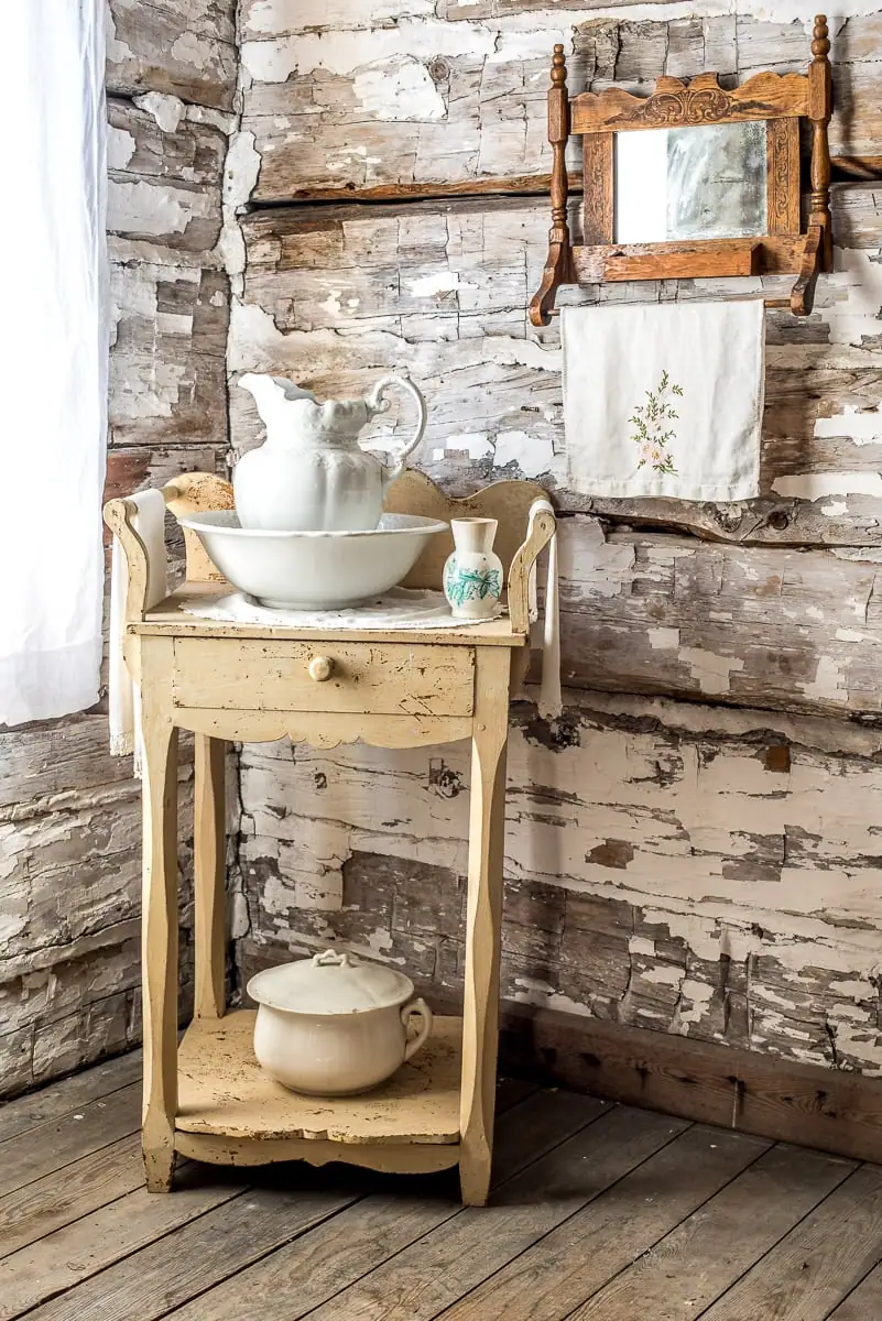 A wooden washstand with white ceramic vessels stands against a weathered log wall beneath a wall mirror.
