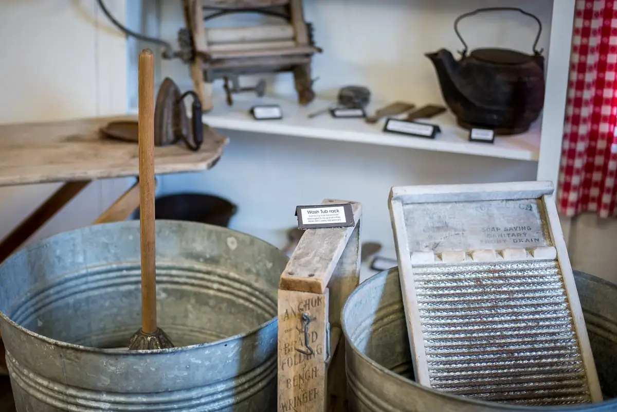 Museum display of galvanized wash tubs, a washboard, a wooden rack, and other household tools.