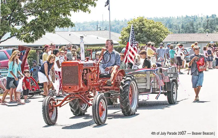 A red vintage tractor pulls a flag-decorated trailer through a crowded street parade.