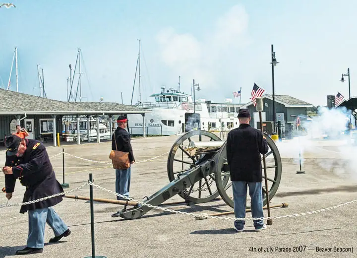 A cannon fires smoke beside a ferry and sailboats as uniformed participants stand behind a chain barrier.