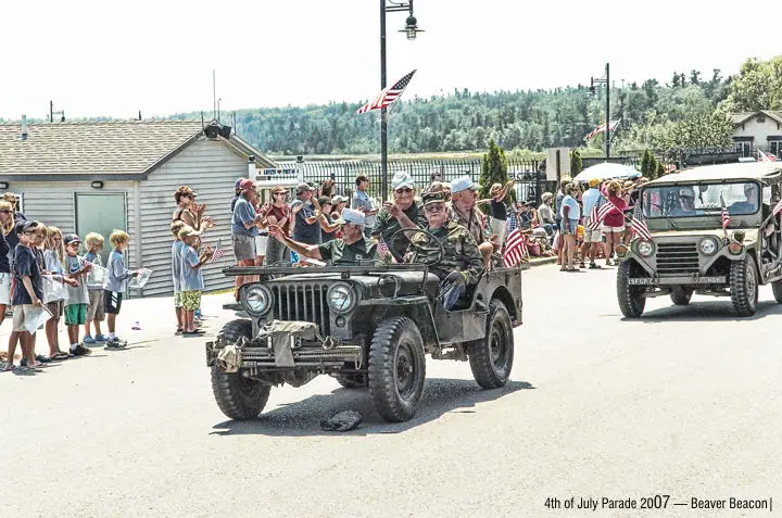 Vintage military jeeps lead a parade past spectators, children, flags, and buildings along a paved street.