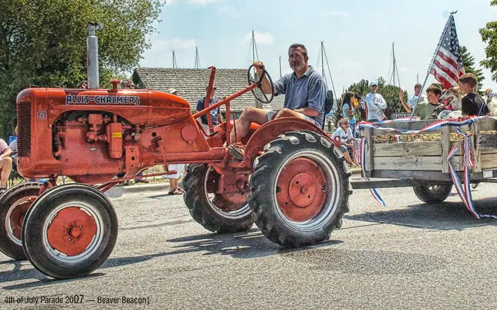 A red tractor pulls a decorated wooden trailer carrying people past spectators and sailboat masts.