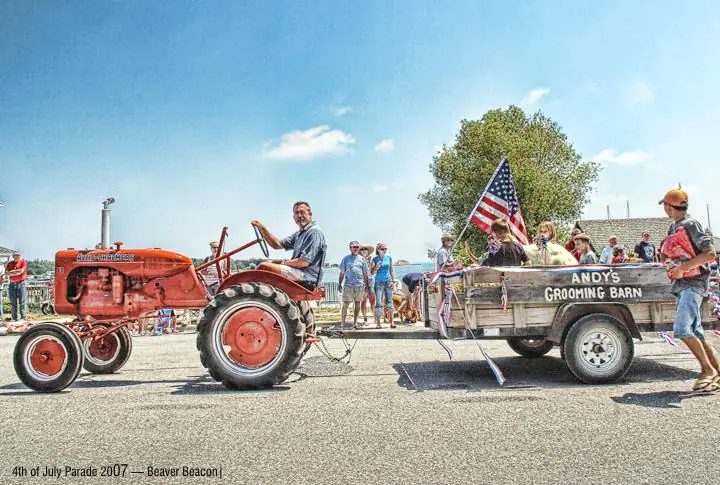 A red tractor pulls a wooden trailer carrying people and an American flag in a street parade.