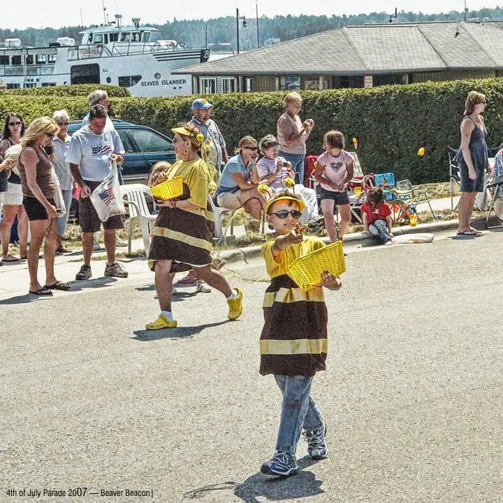 Children in striped bee costumes walk past spectators while a passenger ferry and low buildings appear behind them.