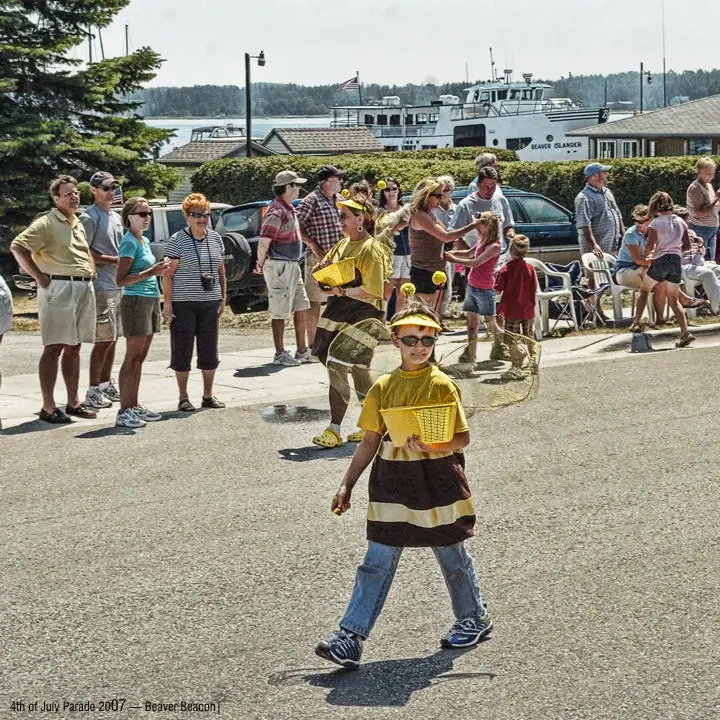 A child in a bee costume walks in front of spectators and a ferry during a parade.