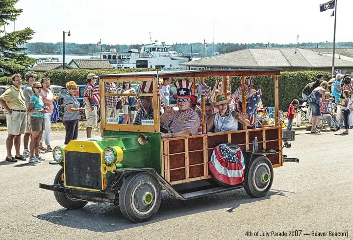A decorated green-and-yellow parade vehicle carries costumed passengers past spectators near the waterfront.