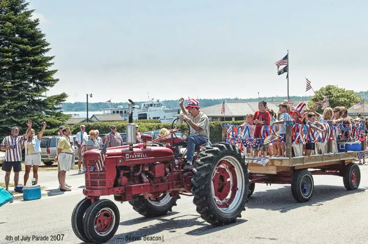 A red tractor pulls a flag-decorated wagon of adults and children past spectators near a ferry.