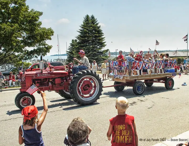 A red tractor pulls a flag-decorated wagon of children past spectators.