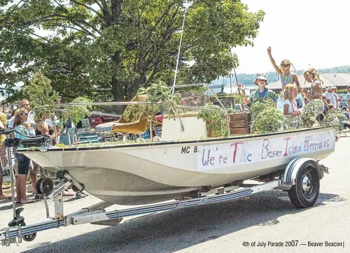 Children and adults ride in a decorated boat trailer with greenery and hand-painted lettering.