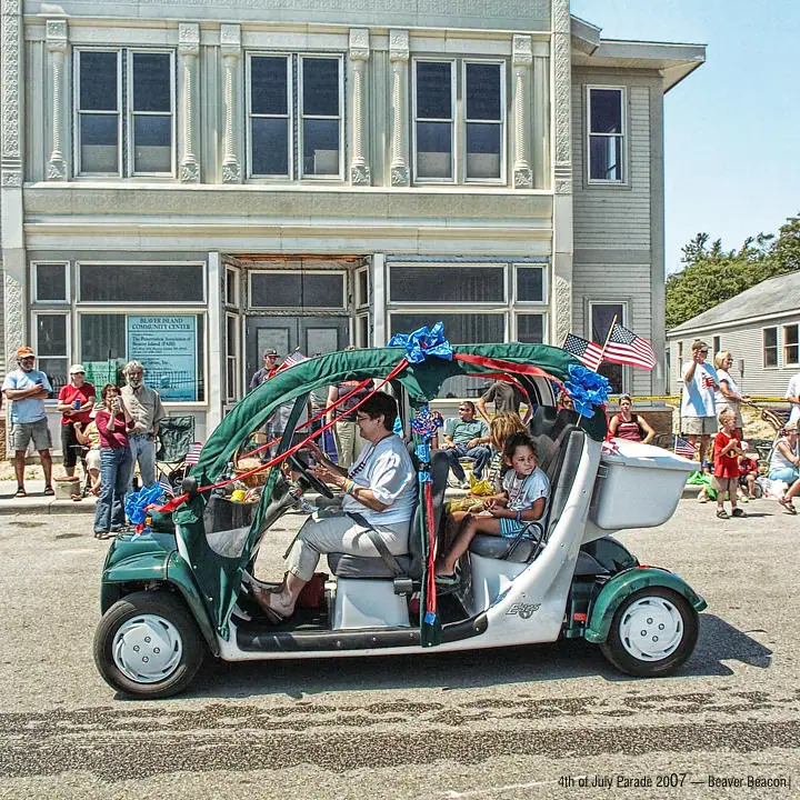 A decorated golf cart carrying a driver and child passes spectators outside an ornate building during a parade.