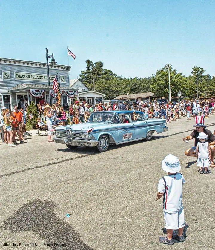 A decorated vintage sedan passes a crowd of spectators and children during a street parade.