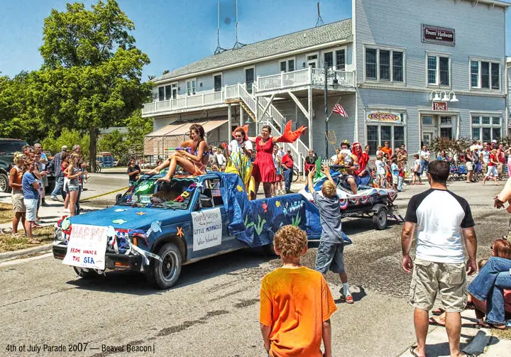 A decorated pickup truck leads a crowded parade past downtown storefronts.