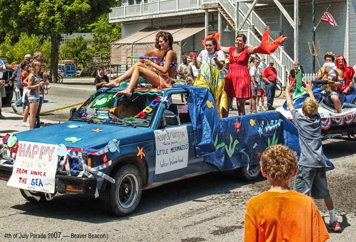 A decorated pickup truck carries a mermaid-themed parade float past spectators and children.