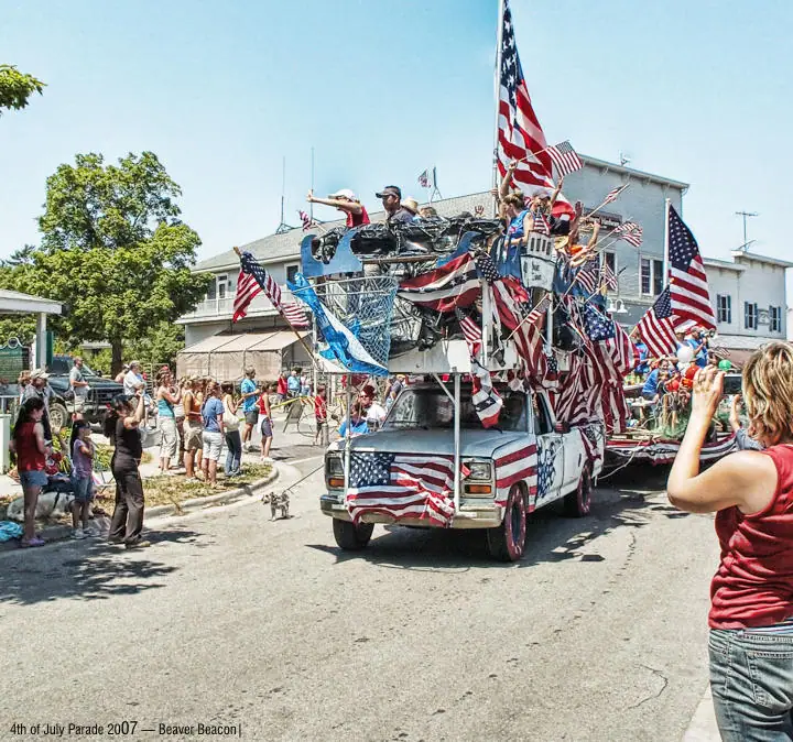 A flag-covered pickup truck float moves along a street as spectators watch and photograph the parade.