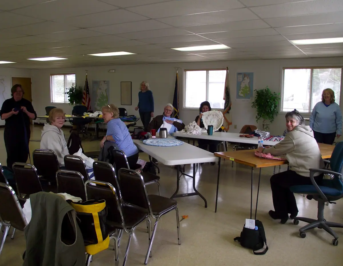 A group of women work on textile crafts around tables in a bright community room.