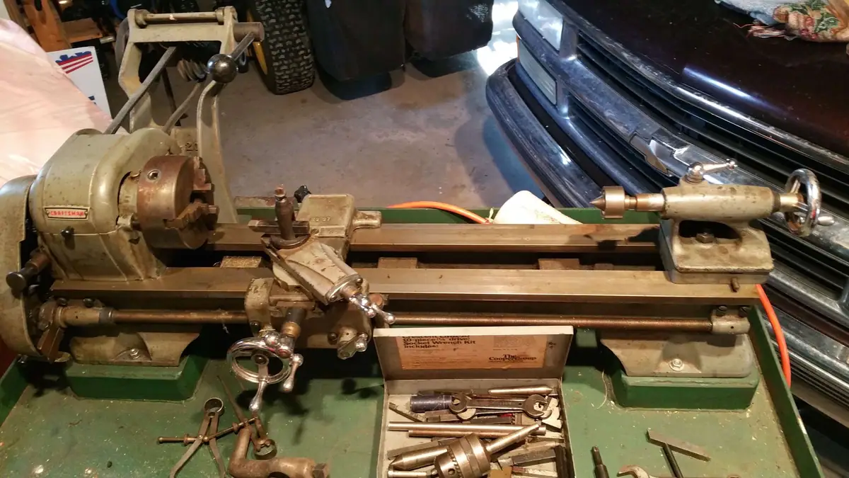 A metal lathe sits on a green workbench beside tools and an open socket set.