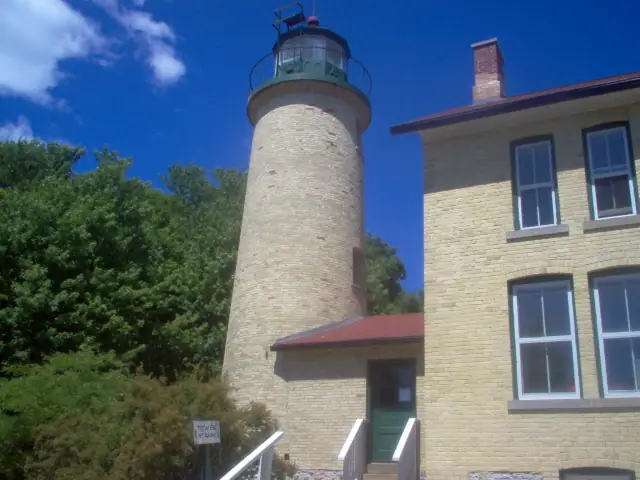 A cylindrical lighthouse tower and adjoining masonry building beside leafy trees under a blue, partly cloudy sky.