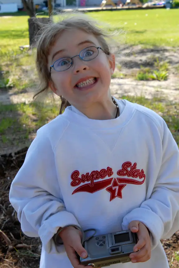 Smiling child in glasses holding a compact digital camera, wearing a sweatshirt marked “Super Star” and “7.”