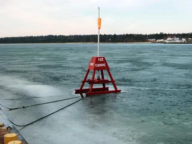 Red buoy marked ICE CLASSIC stands on frozen water beside a mooring line, with a wooded shore beyond.