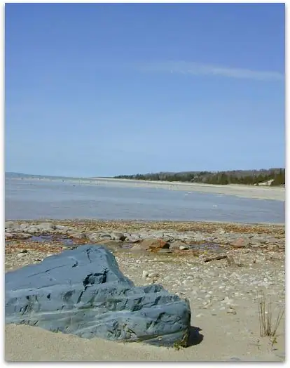 Layered dark rock rests on a pebbly shoreline beside calm water and a distant wooded shore.