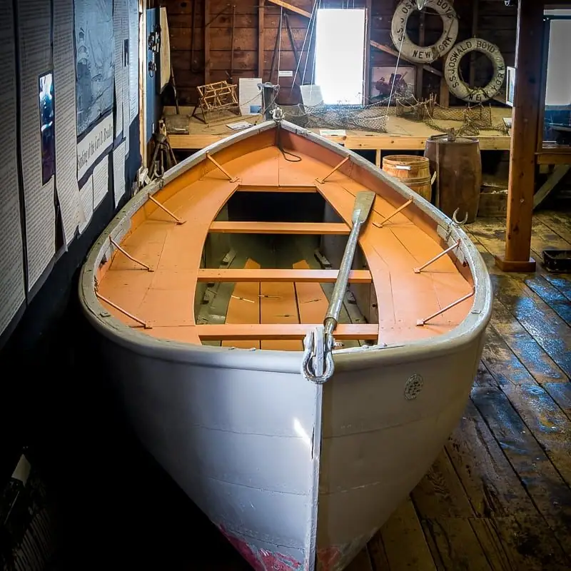 A white and orange rowboat with an oar is displayed indoors beside maritime equipment.
