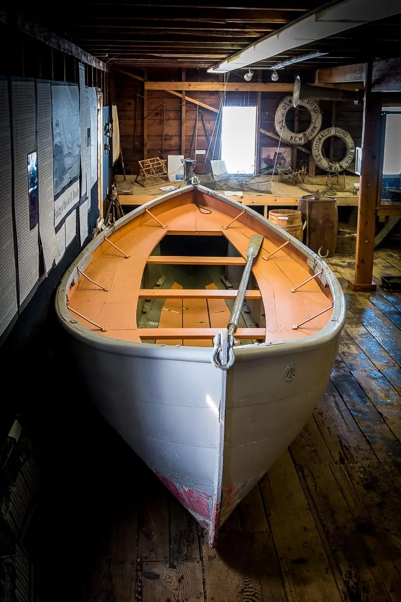 A small open boat displayed indoors beside exhibit panels and two life rings.