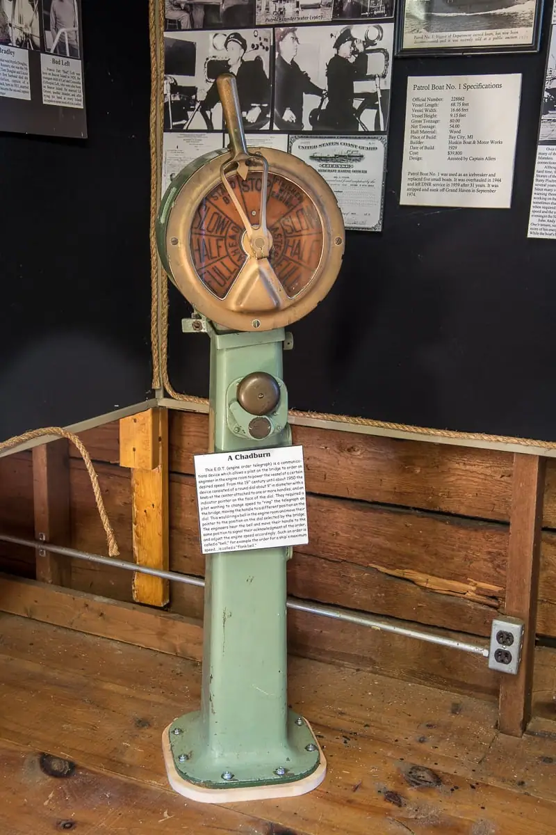A green pedestal-mounted brass marine telegraph stands before a wall of photographs, documents, and exhibit labels.