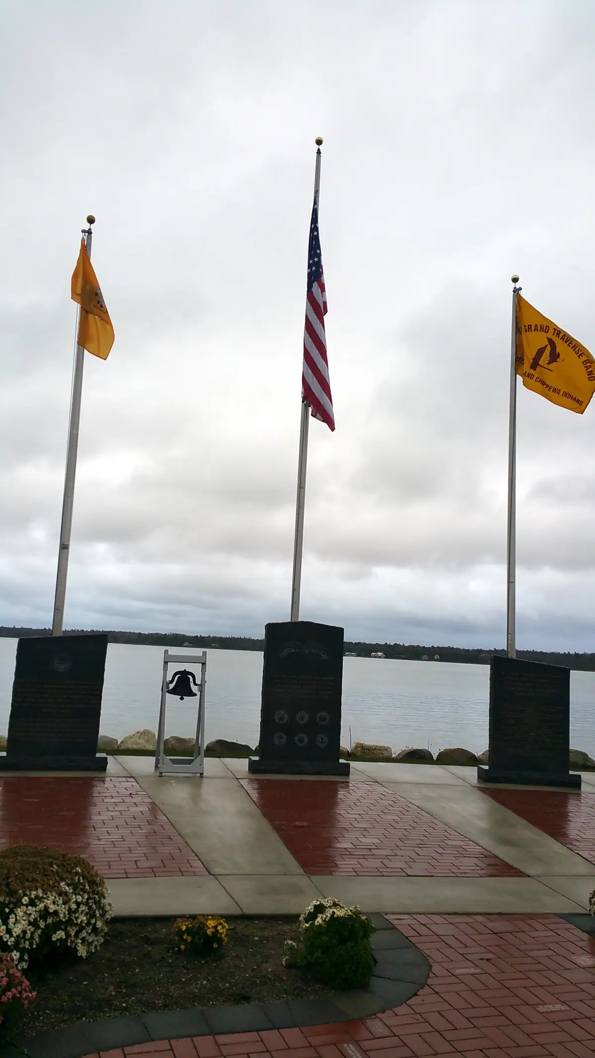 Three flagpoles and dark stone memorials stand beside the water under an overcast sky.