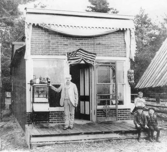 A man and three children stand outside a small brick storefront with an American flag above the doorway.